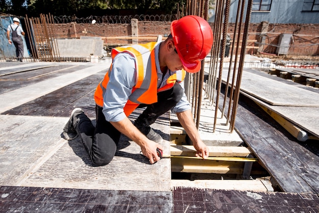 L'uomo vestito con camicia, giubbotto da lavoro arancione e casco misura il foro con un metro a nastro sul cantiere. .