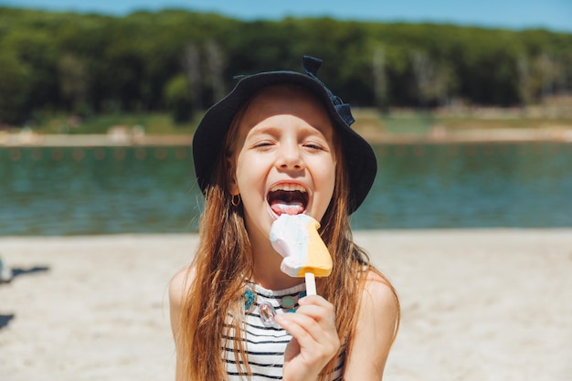 La bambina affascinante con un cappello mangia il gelato sulla spiaggia della spiaggia Concetto di vacanza estiva