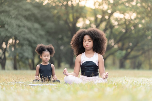 La bambina afroamericana che si siede sulla pratica della stuoia del rotolo medita l'yoga nel parco all'aperto