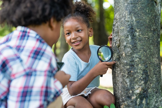 La bambina afroamericana sorridente con gli amici tiene la lente d'ingrandimento per esplorare sull'albero nel parco