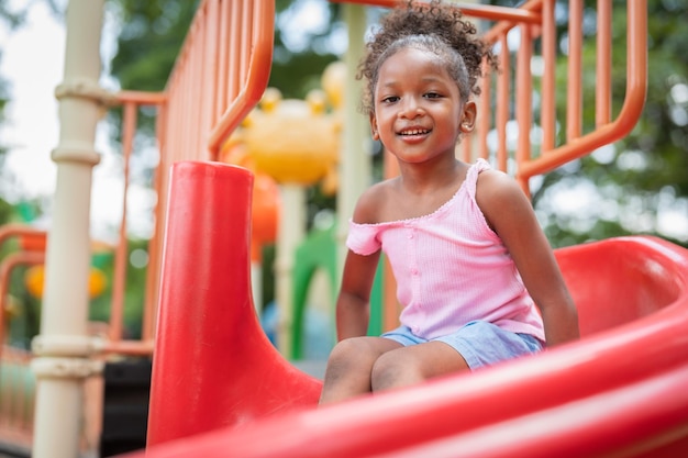 La bambina afroamericana sorridente si diverte a giocare sul dispositivo di scorrimento nel parco giochi nel parco