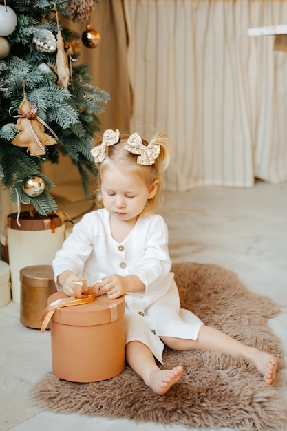La bambina al mattino si siede vicino all'albero di Natale sul tappeto e scioglie il nastro sul regalo di festa. Concetto di vacanza in famiglia