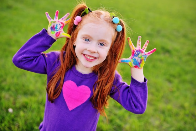 La bambina allegra con capelli rossi mostra le sue mani sporche con vernici multicolori e sorrisi