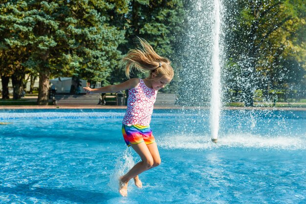 La bambina allegra sveglia sta giocando nella fontana. Il bambino si diverte in un parco estivo nella fontana della città.