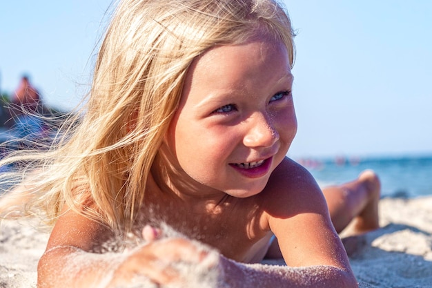 La bambina allegra viene giocata sdraiata sulla spiaggia di sabbia in una giornata di sole.