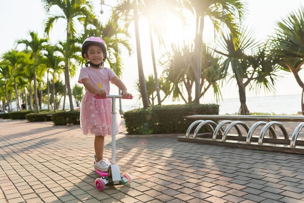 La bambina asiatica felice del bambino indossa il casco sicuro che gioca il bordo di calcio rosa sulla strada nel parco all'aperto il giorno d'estate