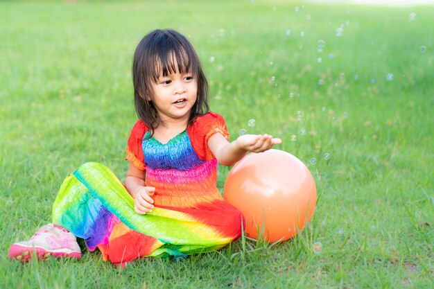 La bambina asiatica porta il vestito variopinto sta giocando le bolle di sapone nel parco