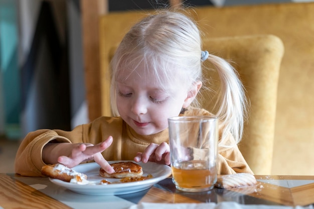 La bambina bionda al caffè al tavolo tocca il dessert con le mani Il bambino al tavolo assaggia i dolci