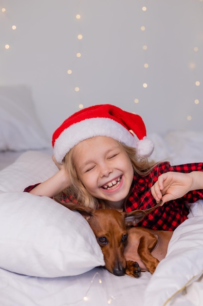 la bambina bionda con un cappello da Babbo Natale e un pigiama dorme a letto con il suo amato cane bassotto