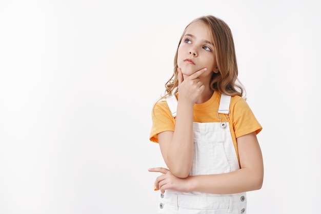 La bambina bionda intelligente e creativa abbastanza giovane guarda pensierosa, tocca il mento facendo la scelta, lo sguardo serio in cima allo spazio della copia, pondera la scelta, risolve il puzzle, stai in piedi sul muro bianco concentrato, pensando