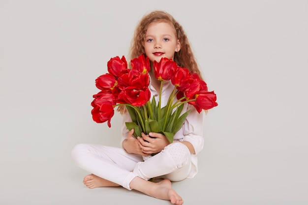 La bambina bionda seduta sul pavimento con le gambe incrociate e tenendo il mazzo di tulipani rossi, guardando la parte anteriore, ha un regalo piacevole