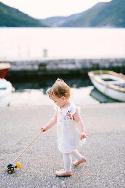 La bambina cammina lungo il molo con un giocattolo sullo sfondo di montagne marine e barche parcheggiate