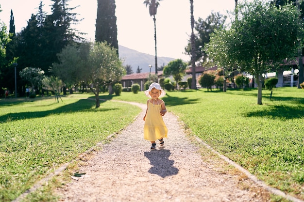 La bambina cammina lungo un sentiero di ghiaia in un parco verde sotto il sole splendente