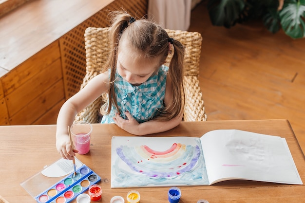 La bambina carina disegna con un pennello e dipinge al tavolo in soggiorno