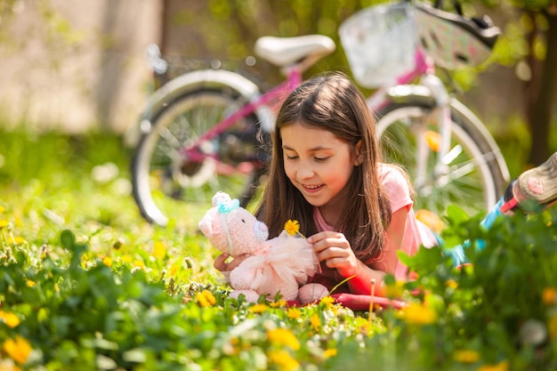 La bambina carina è sdraiata su un prato verde e gioca con un orsacchiotto rosa lavorato a maglia e un dente di leone La sua bicicletta rosa sullo sfondo
