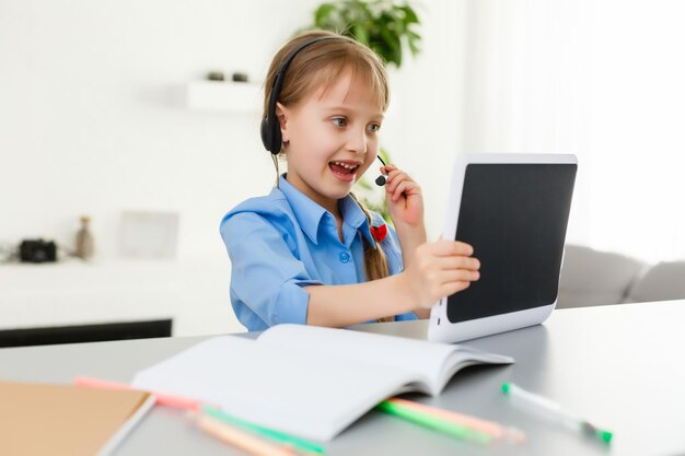 La bambina carina è seduta a tavola con il suo laptop e studia online