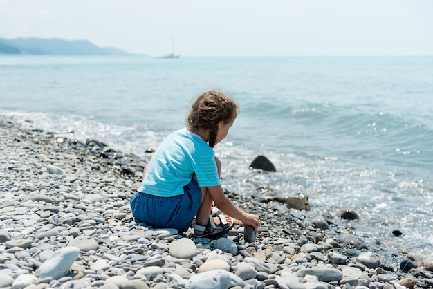 La bambina carina si siede su una spiaggia di ciottoli e gioca con i ciottoli