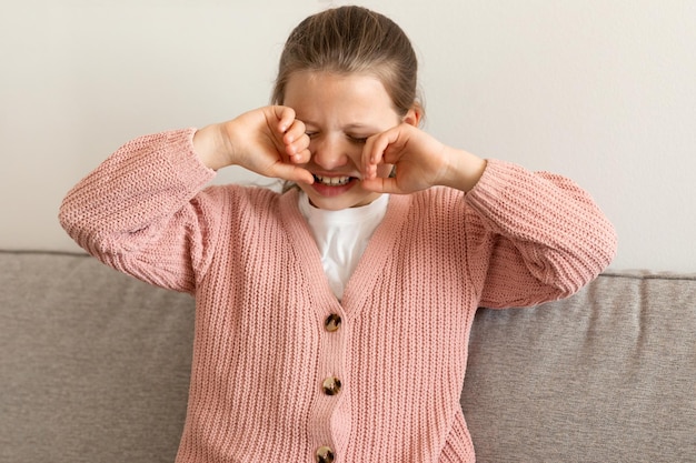 La bambina caucasica piangente triste infelice pulisce il viso con le mani si siede sul divano nel soggiorno