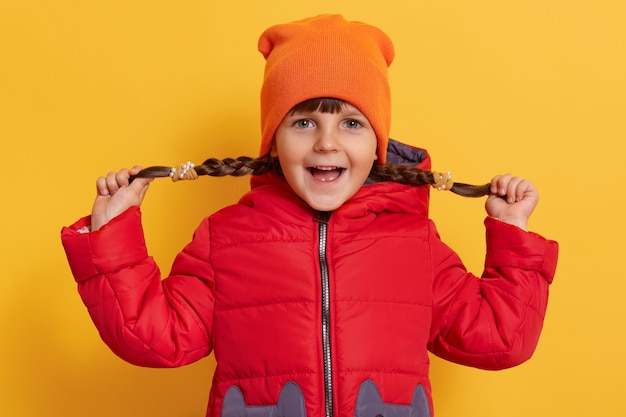 La bambina che tira le sue trecce ai lati, guardando davanti con l'espressione facciale eccitata, tiene la bocca aperta, indossa abiti caldi, in piedi isolato sopra il muro giallo