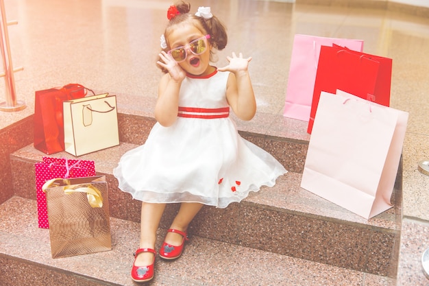 La bambina con gli occhiali da sole si siede sui gradini del centro commerciale con lo shopping