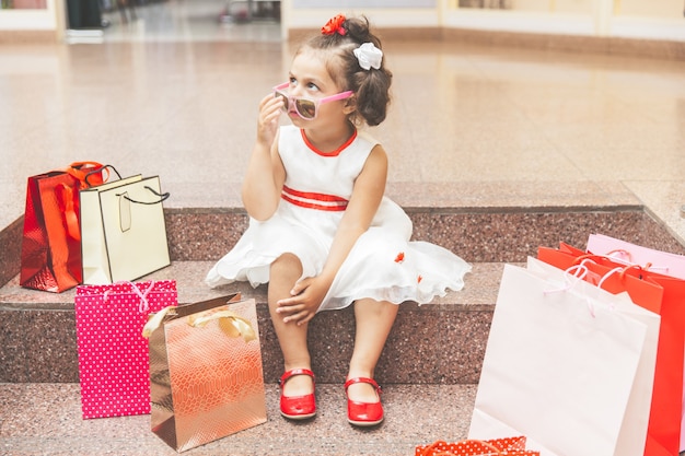 La bambina con gli occhiali da sole si siede sui gradini del centro commerciale con lo shopping