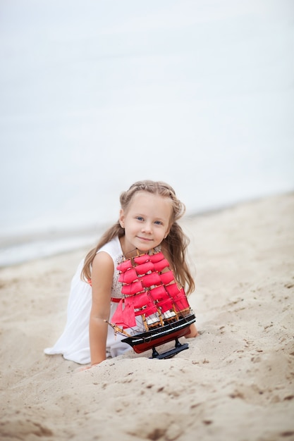 La bambina con i capelli biondi in abito bianco si siede sulla spiaggia e tiene in mano una nave giocattolo con vele rosse