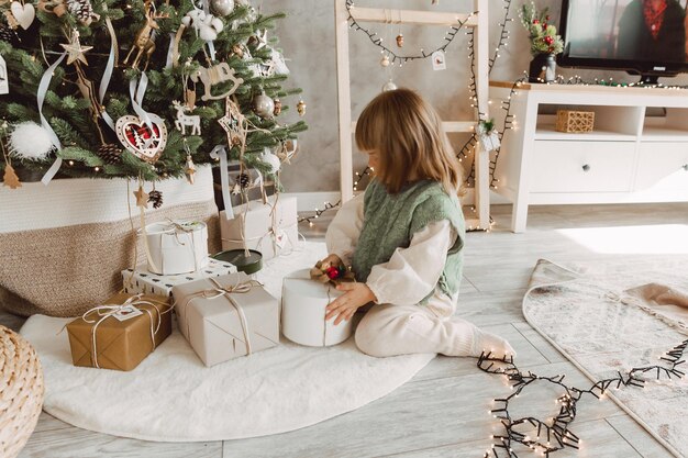 La bambina con i capelli biondi si siede sul pavimento vicino all'albero di Natale con i regali.