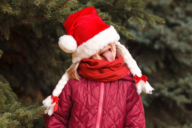 La bambina con il cappello di Natale se lo tirò sugli occhi e tirò fuori la lingua Bambina divertente di Capodanno