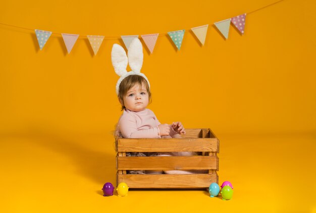 La bambina con le orecchie di coniglio si siede in una scatola di legno con uova colorate su uno sfondo giallo