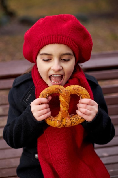 La bambina con un berretto rosso e un cappotto si siede su una panchina e mangia un pretzel. Foto di alta qualità