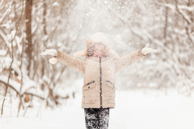 La bambina con un cappello bianco e una giacca nella foresta invernale lancia la neve. Divertimento invernale