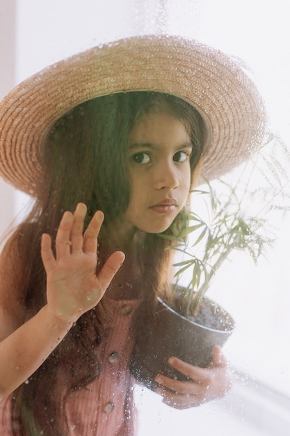 la bambina con un fiore al coperto in vaso guarda fuori dalla finestra attraverso il vetro bagnato dalla pioggia