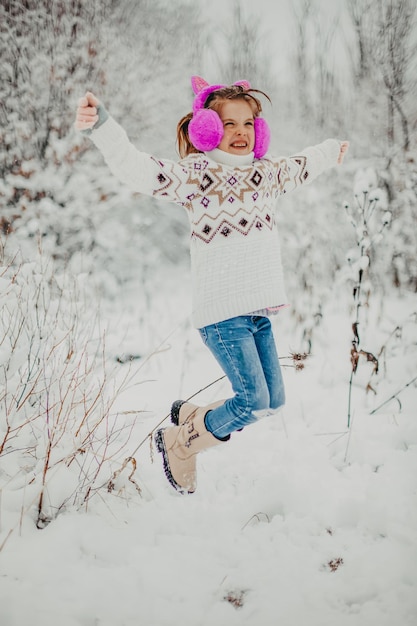 La bambina con un maglione e le cuffie invernali salta e si diverte in una giornata invernale.