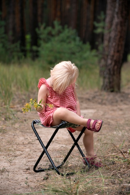 La bambina con un mazzo di fiori di campo abbassò imbarazzata la testa. Il bambino sta riposando sulla sedia pieghevole nella foresta. Cornice verticale.