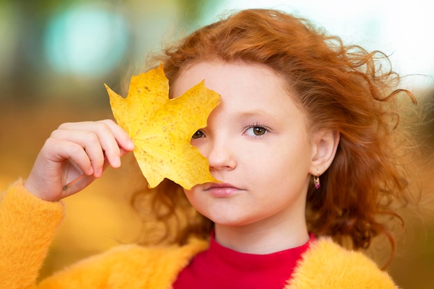 La bambina dai capelli rossi chiude un occhio con una foglia d'acero gialla Bambino triste in un primo piano del giorno d'autunno