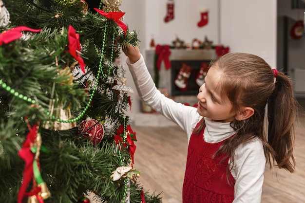 La bambina decora l'albero di Natale Bambino in abiti natalizi guarda l'albero di Natale