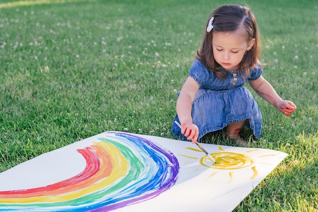 La bambina di 2-4 anni dipinge arcobaleno e sole su un grande foglio di carta, seduta su un prato verde