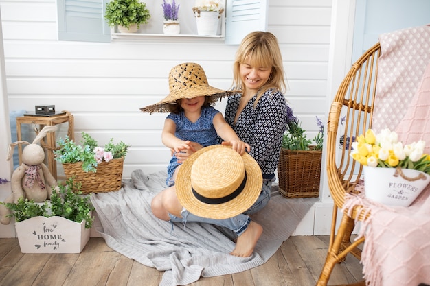 La bambina di 4-5 anni sta giocando con i cappelli di paglia sulla veranda di casa con la madre in braccio. Risate, felicità, gioia, concetto di maternità