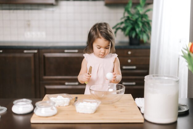 La bambina di due anni sta imparando a cuocere la torta in piedi rompendo le uova