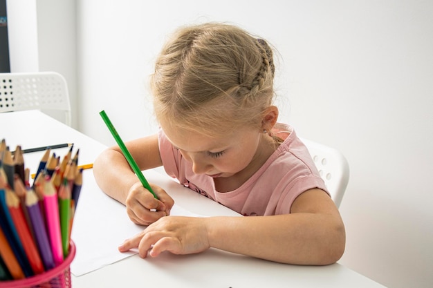 La bambina disegna con matite colorate seduta a un tavolo bianco su sfondo bianco