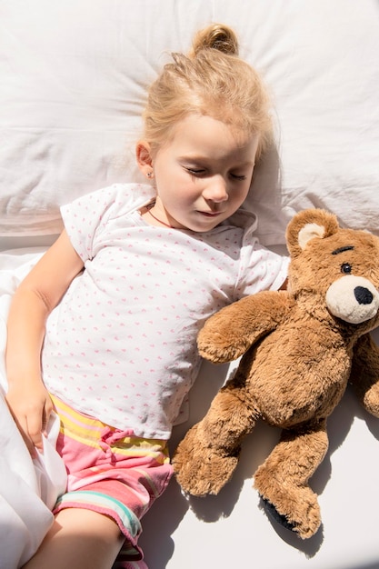 La bambina dorme abbracciando un orso giocattolo morbido su un letto bianco Vista dall'alto piatto