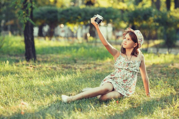 La bambina è giocata dalla macchina fotografica seduta sull'erba nel parco. Fare selfie e fotografare il mondo intorno