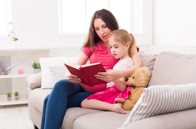 La bambina e il suo libro di lettura della mamma seduti sul divano di casa. Madre e figlia studiano le fiabe. Maternità, attività e interessi comuni, sviluppo precoce, concetto di cura, spazio di copia