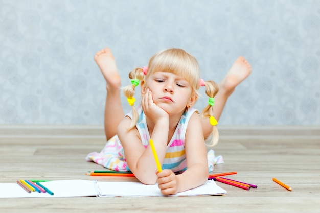 La bambina felice bionda disegna sdraiata sul pavimento a casa. Educazione della prima infanzia