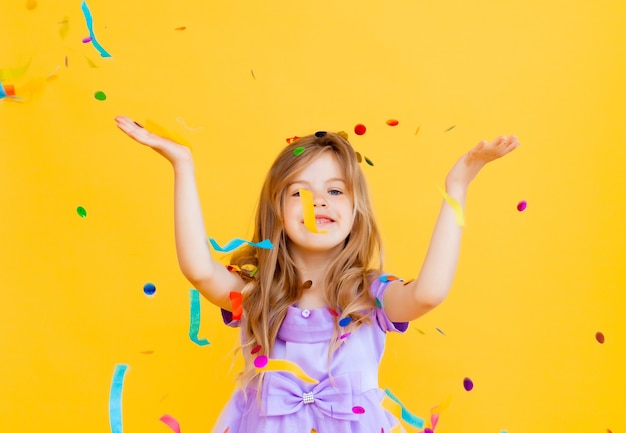 La bambina felice con i capelli biondi e in un vestito blu cattura i coriandoli su uno sfondo giallo, concetto di vacanza