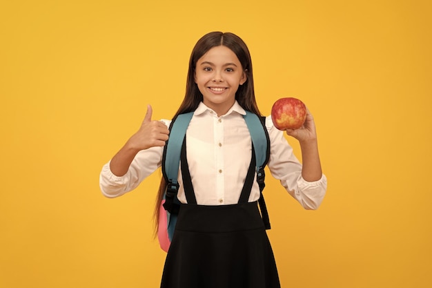 La bambina felice dà i pollici in su che tiene l'istruzione gialla del fondo della borsa di scuola e della mela