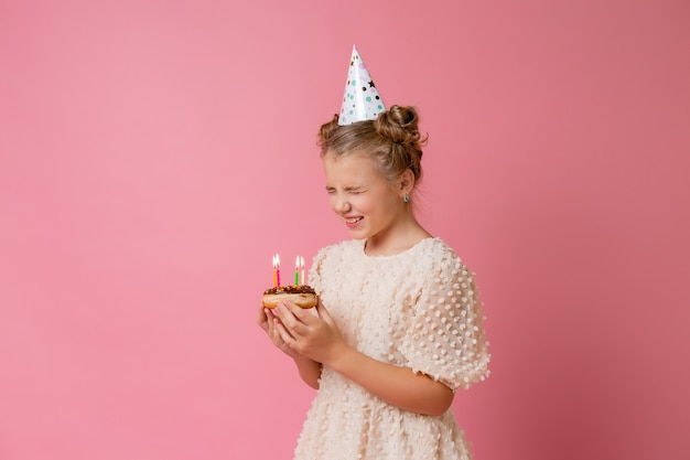 La bambina felice in un berretto per il suo compleanno esprime un desiderio e spegne le candeline su una torta.