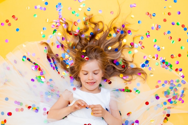 La bambina felice in un vestito bianco celebra una festa con vista dall'alto di coriandoli. Bambino di compleanno, emozioni positive.