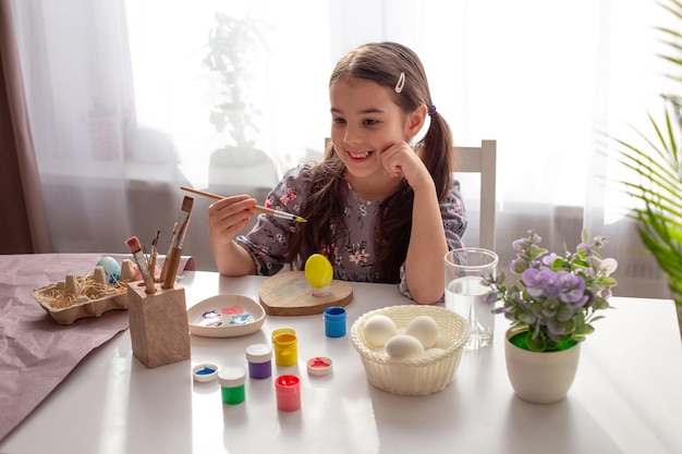 La bambina felice si siede a un tavolo bianco vicino alla finestra tiene un pennello un uovo da colorare