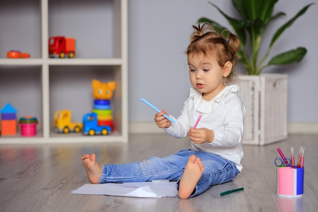 La bambina felice si siede sul pavimento e disegna su carta con matite colorate